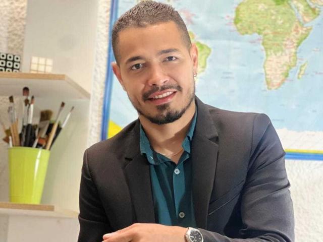 AlexCarls, short-haired dude with a beard, black suit, teal shirt, smiling, holding arms, map in background, paintbrushes in green cup on left, small black cube on shelf.
