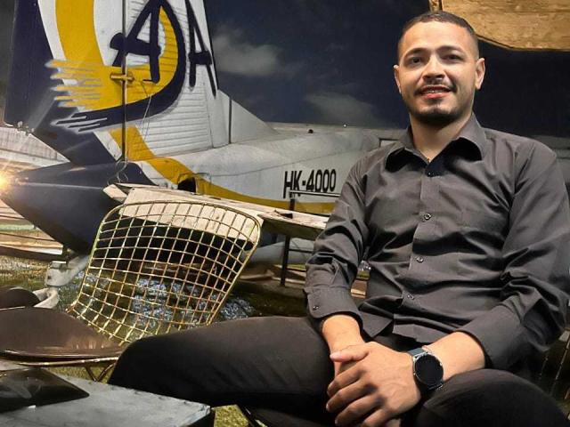 AlexCarls, with a short beard and black shirt, sits in front of a vintage airplane with a yellow and blue logo, wearing a black watch, smiling.