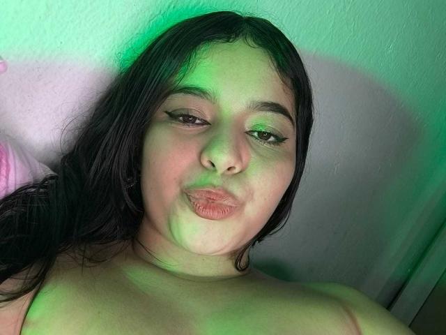 LissyCarter with puffy lips, green light on her face, black wavy hair, and eyeliner, looking at the camera, topless, showing her bare shoulders.
