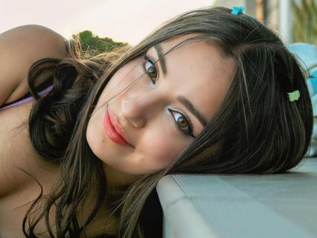 IsadoraGallego with dark brown hair, green eyes, and pink lips, lying down with a small blue and green hair clip, smiling softly, wearing a purple top, with a relaxed look.