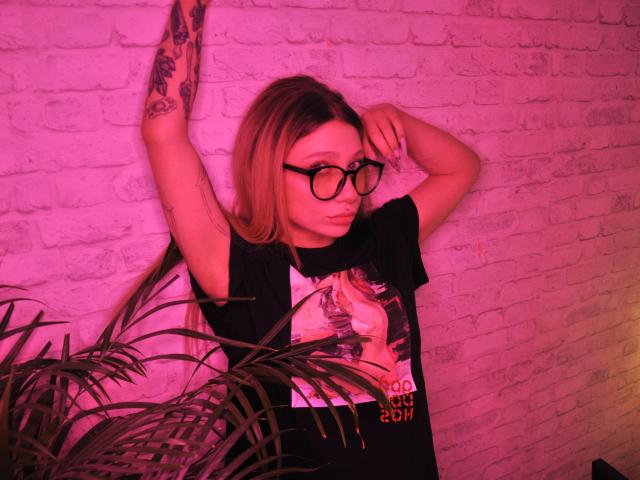 AliceAndCristal with long blonde hair, black glasses, and tattoos, wearing a black shirt, arms raised, pouty face, pink lighting, and a plant in the foreground.