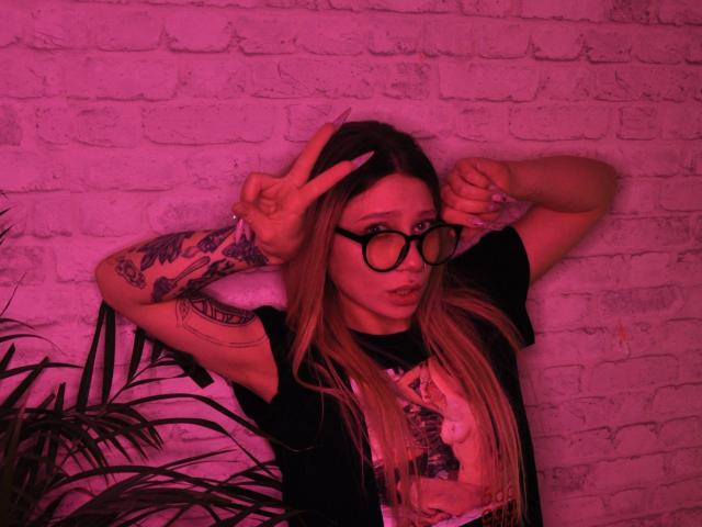 AliceAndCristal with long blonde hair, black glasses, and tattoos, making a peace sign and rock sign with pink neon lighting, wearing a black shirt with a white graphic.
