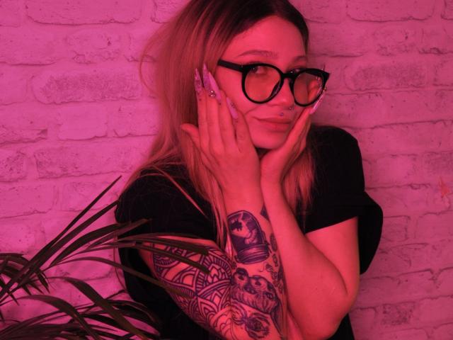 AliceAndCristal with glasses, blonde hair, black shirt, tattooed arm, hands on face, pink light, plant in foreground, smiling subtly, looking cute, red-tinted skin.