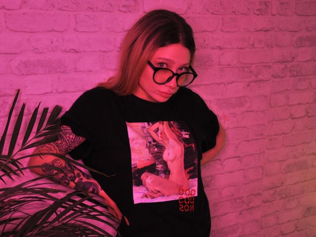 AliceAndCristal with pink-tinted hair, wearing round glasses, black t-shirt with graphic, tattooed arm, standing next to potted plant, looking sideways, pink light, white brick wall.