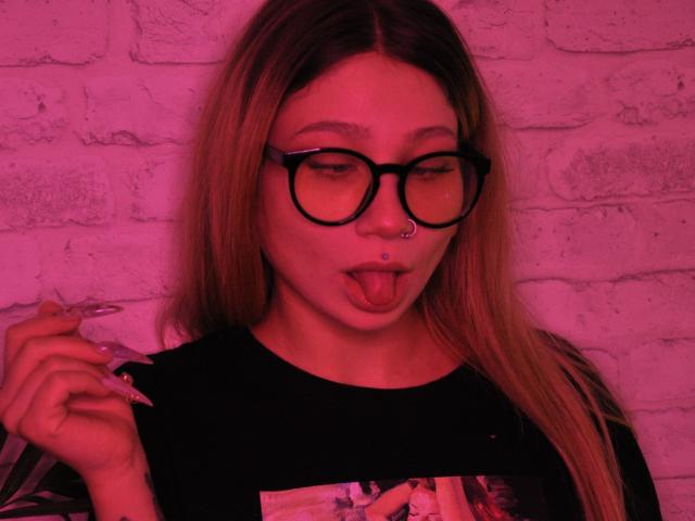 AliceAndCristal with long blonde hair, black glasses, nose ring, and tongue out, holding a cigarette, wearing a black shirt, illuminated by pink neon light.