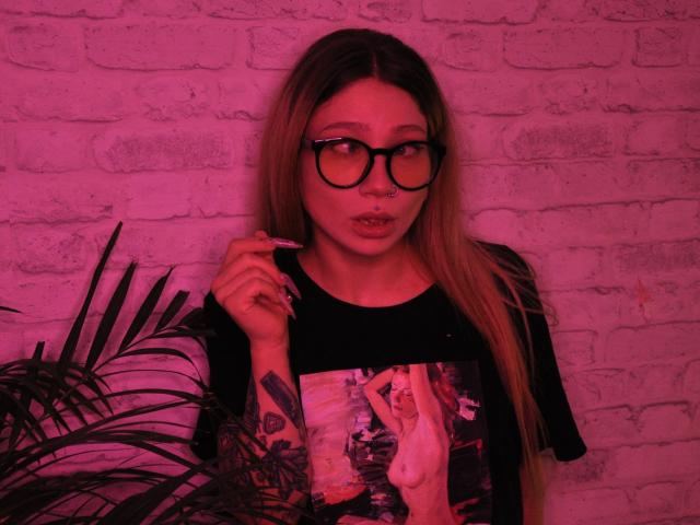 AliceAndCristal with pink-tinted glasses, long blonde hair, black shirt with naked woman print, holding nail, tattoo on right arm, pensive expression, leafy plant on left.