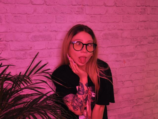 AliceAndCristal with long blonde hair, black glasses, and tattooed arm, sticking out tongue, hand near face, wearing black shirt, pink-hued, with green plant on left.