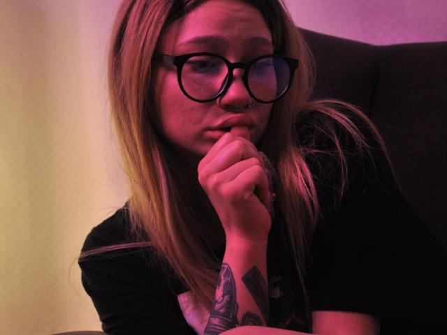 AliceAndCristal with blonde hair, black glasses, nose ring, and tattooed arm, biting her finger, looking serious in a pinkish-purple light. Wearing a black shirt.