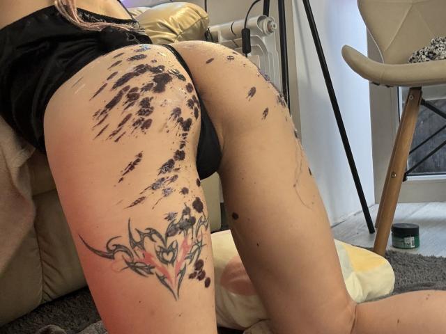 AliceAndCristal's ass is covered in black paint splatters, showing a tattoo on her right thigh, wearing black panties, on a beige couch, with a chair in the background.