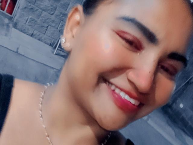 SusanaDiaz with medium brown skin, dark eyebrows, red eyeliner, pink lipstick, smiling, nose piercing, small earrings, chain necklace, black top, slightly blurry selfie.