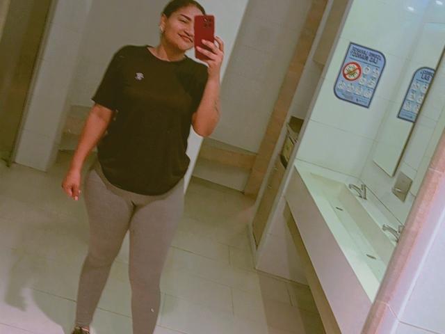 SusanaDiaz with a smile, black short-sleeve shirt, gray tight leggings, holding a red phone, standing in a bathroom, showing a curvy, sexy, and confident ass.