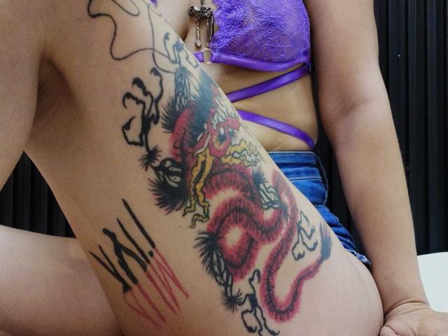 Close-up of LolyYunno's side, showing a vibrant, detailed tattoo of a red and black dragon with yellow eyes and white flames. The dragon's body is coiled, surrounded by black, spiked lines and smaller black tattoos. LolyYunno wears a purple lace bra and blue denim shorts. The tattoo's bold lines and bright colors contrast with the surrounding skin. The dragon's textured scales and fiery accents add depth to the design. LolyYunno's tan skin and the tattoo's vivid colors create a striking visual.