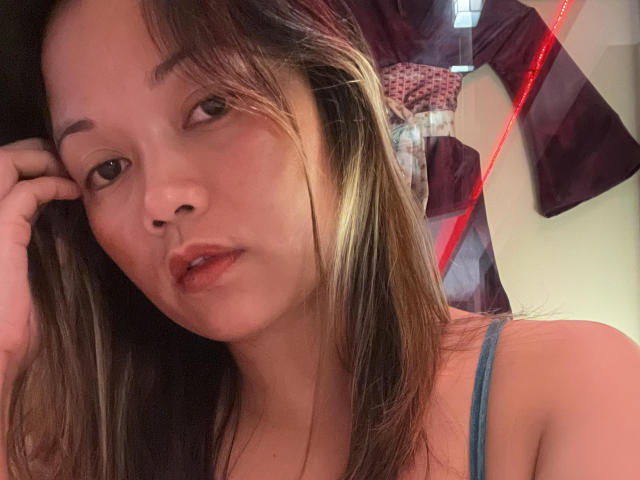 AsianBoobGoddess with straight brown hair, subtle highlights, and light skin, wearing a blue spaghetti strap, looking at the camera with slightly parted red lips, in a dimly lit room.