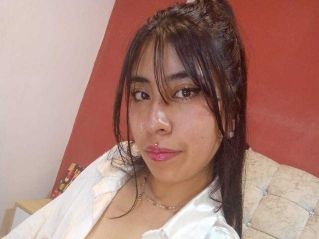 LinaSpearce has medium brown skin, black hair in a messy ponytail, brown eyes, pink lipstick, a shiny nose, and wears a white shirt with a pearl necklace.