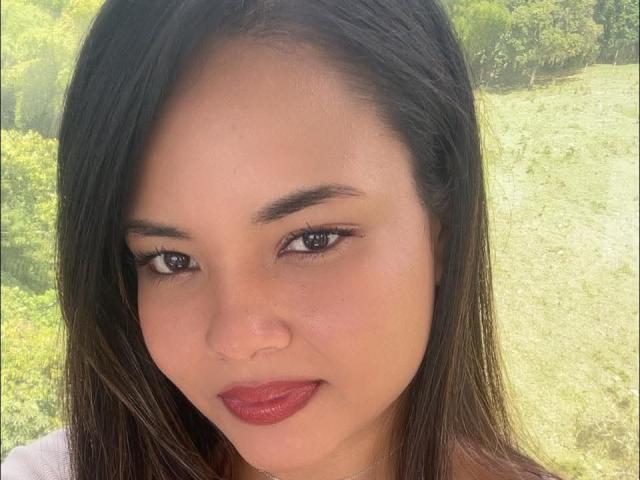 LuckyMillie has medium brown skin, dark brown eyes, and red lipstick. She has straight black hair and a serious expression. Her tits are not visible.