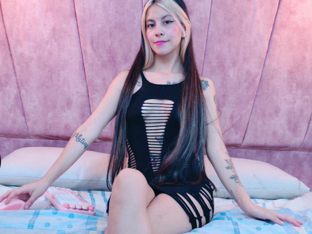 Yliria with long black and blonde hair, wearing a black cut-out dress, sitting on a bed, showing tattoos on her arms, pink lipstick, and a cute smile.