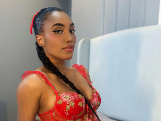 ChelseaDow with dark skin, red bra with gold leaf patterns, long black braided ponytail, red hair ribbon, sitting on bed, looking at camera, red lips, subtle makeup.