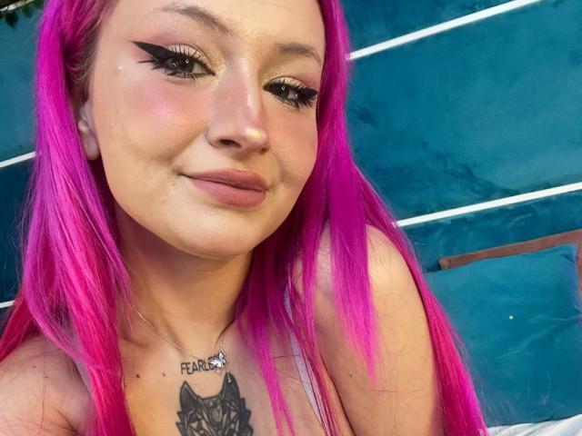 SunnyGlow with bright pink hair, heavy black eyeliner, subtle pink lipstick, and a cat tattoo on her chest, smiling softly. She's wearing a silver necklace that says "FEARLESS."