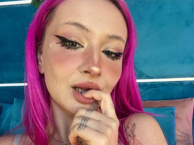 SunnyGlow with bright pink hair, biting her finger, showing off thick black eyeliner and glossy pink lips. Her hand has tattoos and she's wearing a pink top.