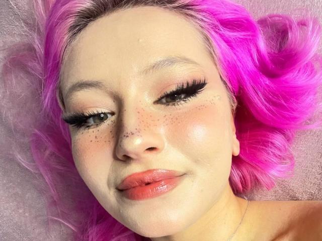 SunnyGlow has pink hair, freckles, and glossy pink lips. She's lying down with her eyes half-closed, showing long black eyelashes. Her skin is shiny and she's smiling.