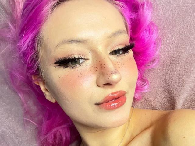 SunnyGlow with pink hair, freckled face, glossy pink lips, and long black eyelashes, looking slightly to the side, showing off her cute, sexy face.