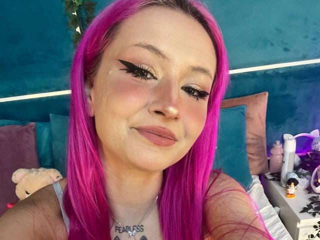 SunnyGlow with bright pink hair, black eyeliner, and pink lipstick, smiling at the camera. She has a nose ring and a necklace with "FAIRIES" in cursive.