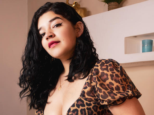 NaomiCotton with wavy black hair, wearing a leopard print top, showing her tits, red lipstick, and a gold necklace, smiling at the camera.