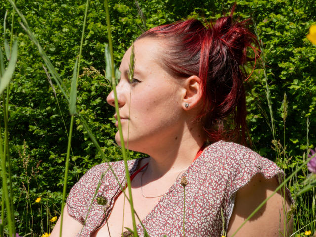 PimentMaya has red-streaked hair in a messy bun, facing left. She wears a short-sleeved, floral-patterned top with a red strap visible. Her skin is fair with a few small moles. She has a small stud earring in her left ear. Tall green grasses partially obscure her. Bright green foliage forms the background. The light is natural, highlighting her profile and the textures of her hair and top. The image focuses on her upper body and face. Pictorial realism with a casual, everyday feel.