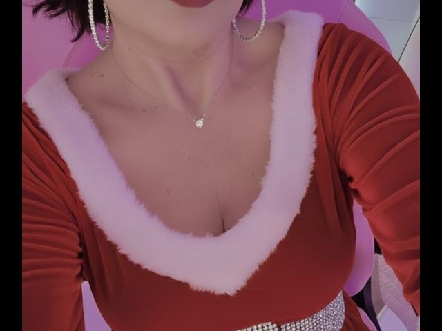 Cateys69's red velvet top with pink fur trim, showing her cleavage and a diamond necklace, wearing large hoop earrings, and a sparkling belt, with red lipstick.