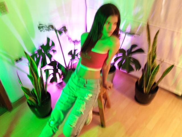 EmyCarther with long dark hair, red crop top, and ripped jeans, leaning on a chair, green and purple lights, looking back, small tattoo on her right side.