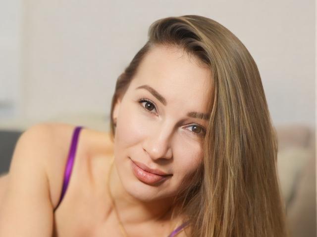 BrandyStone has light brown hair, a cute face, and is wearing a purple bra, smiling softly at the camera with brown eyes and a slight tilt of her head.