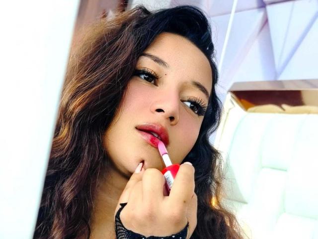 NicoleMartins with wavy brown hair, holding a red-and-white lipstick to her slightly parted red lips, looking at the camera with half-closed eyes, wearing black lace.