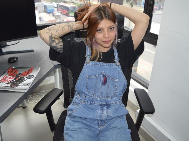 SaraxNavyx, with arms up, wearing blue denim overalls, black shirt, and tattoos, sits in a chair, looking at camera, brown hair, slightly smiling.