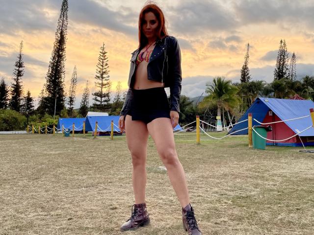 Ariss69 with red hair, black leather jacket, red bra, black shorts, and boots, standing on grassy field, hands by sides, sunset in background, tall trees and colorful tents.