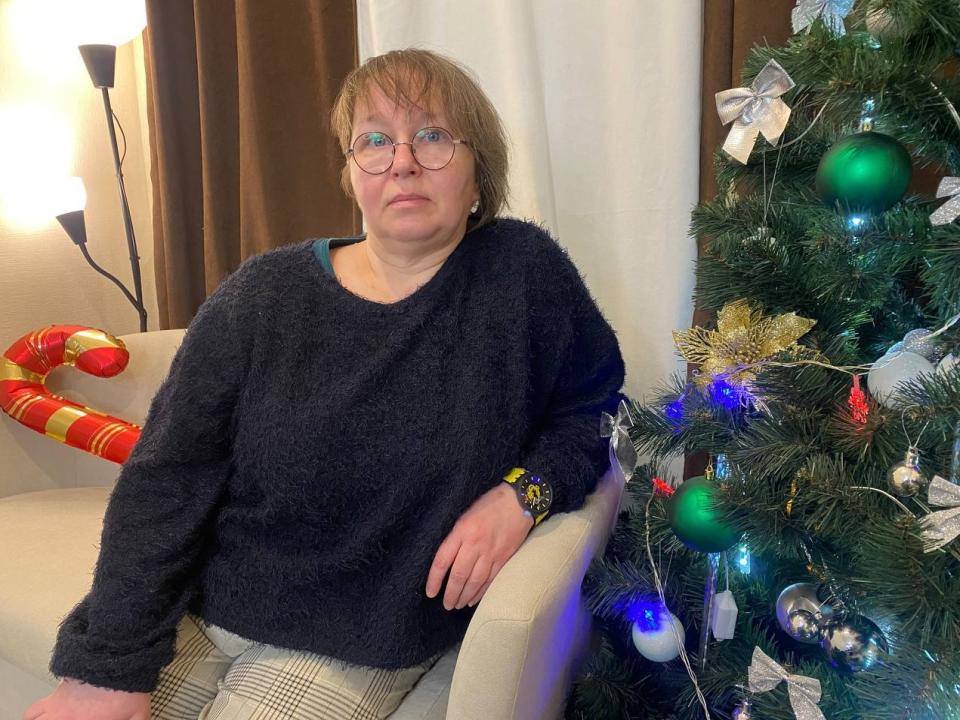 TinaBlush, middle-aged woman with short brown hair, wearing glasses and a dark blue fuzzy sweater, sits next to a decorated Christmas tree with green and blue ornaments.
