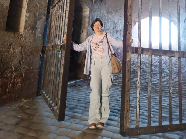 LunaDobra with short black hair, light pink shirt, blue pants, and brown shoes, standing in a cobblestone hallway, holding a metal gate with both hands, smiling.