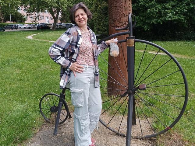 LunaDobra stands with a bicycle wheel sculpture, wearing a plaid shirt, pink floral top, white cargo pants, and pink shoes, smiling with one hand on her hip.