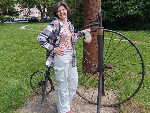 LunaDobra stands with hand on hip, wearing a plaid shirt, white pants, and pink shoes, holding a bike wheel sculpture. Grass and trees in background. Smiling.