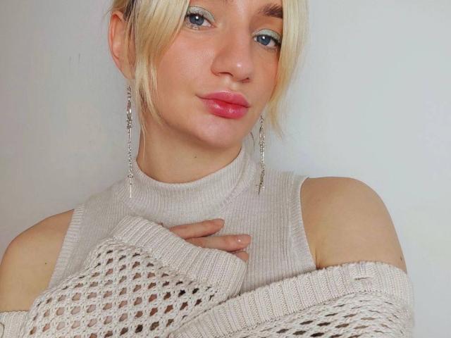 Arina, con i capelli biondi, gli occhi azzurri e le labbra rosa, indossa un maglione bianco a maglia, con un motivo a rete e con le spalle scoperte, e orecchini lunghi e pendenti che le sfiorano il petto..