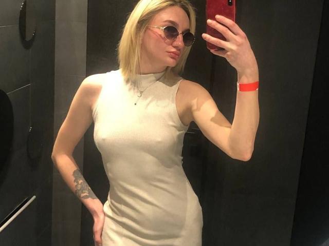 Arina, gekleed in een strakke witte jurk, die haar kleine borsten laat zien en een tatoeage op haar rechterarm heeft, maakt een spiegelzelfportret met een rode telefoon, draagt een bril en een rode polsband..