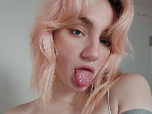 ArinaWinss with pinkish blonde hair, sticking out a wet tongue, blue eyes, light skin, and a silver necklace, wearing a thin-strapped top, showing a subtle off-shoulder look.