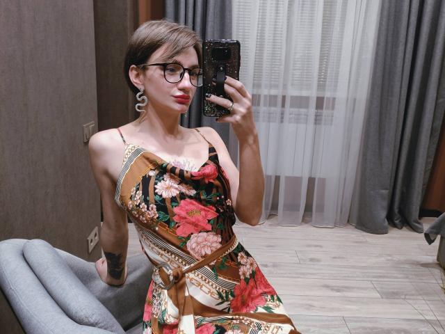 Arina, con i capelli castani corti, gli occhiali e il rossetto rosso, indossa un abito floreale, tiene in mano un telefono e mostra un tatuaggio sul braccio destro, seduta su una sedia grigia..
