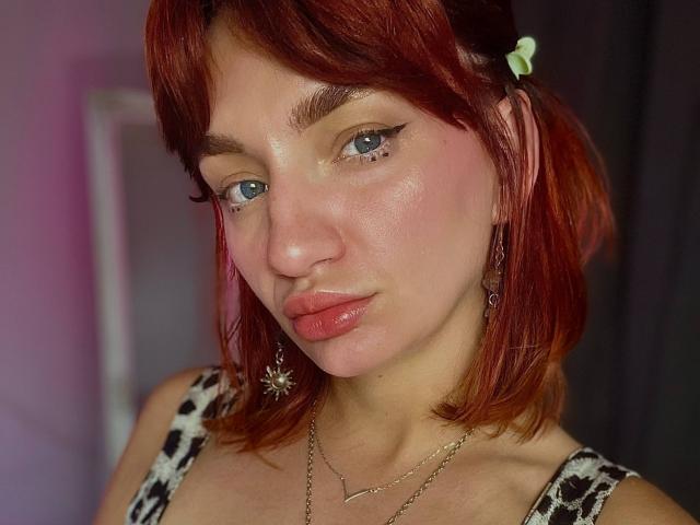ArinaWinss tiene el pelo rojo, ojos azules y labios rosados y brillantes. Lleva una blusa con estampado de leopardo y aretes con forma de estrella, con una pinza para el pelo verde discreta. Su rostro está ligeramente sudoroso..