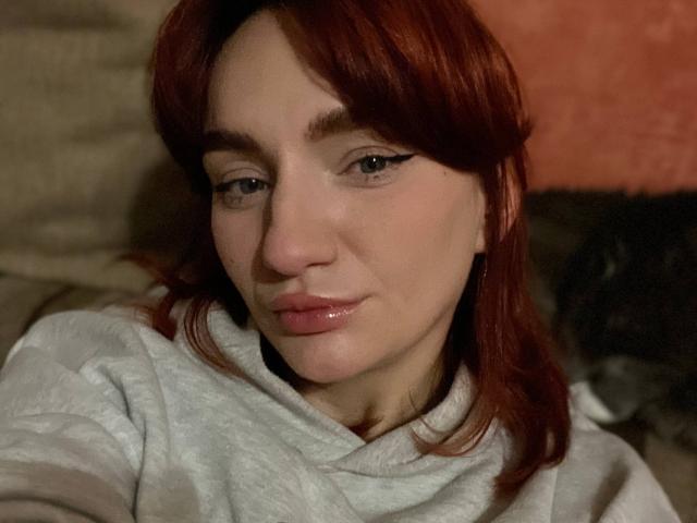 Arina, met rood haar, lichte huid en roze lippen, draagt een grijze hoodie, heeft subtiele eyeliner en een lichte glimlach, en kijkt naar beneden met een ontspannen uitdrukking..