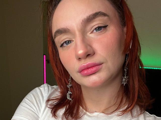 Arina, con cabello rojo, camisa blanca, labios rosados, delineador de ojos y aretes de mariposa, mirando ligeramente hacia un lado con una sonrisa sutil, mostrando su rostro ligeramente sonrojado y adorable..