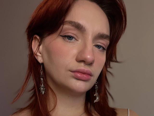 ArinaWinss has red hair, light skin, and pink lips with subtle eyeliner. She's wearing dangling earrings with small white stones. Her expression is neutral and slightly pouty.