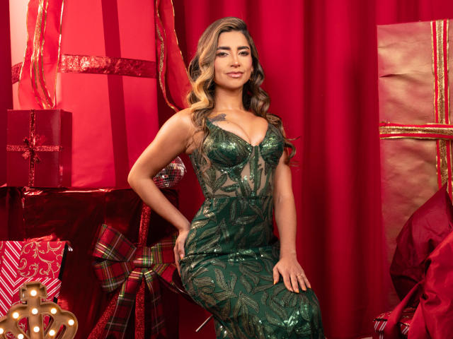 SofyaLane, con cabello castaño ondulado, vestida con un vestido verde ajustado adornado con lentejuelas, que resalta sus pechos y glúteos, está de pie frente a regalos envueltos en papel rojo y dorado..