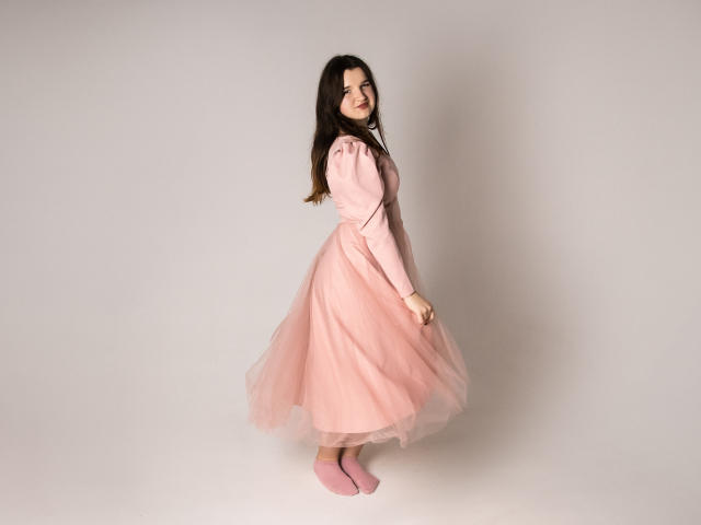 BonnieFlower with long brown hair, pink puffed sleeves dress, and pink tulle skirt, standing sideways, smiling, pink socks, soft light, gentle pose, cute, fluffy, feminine.