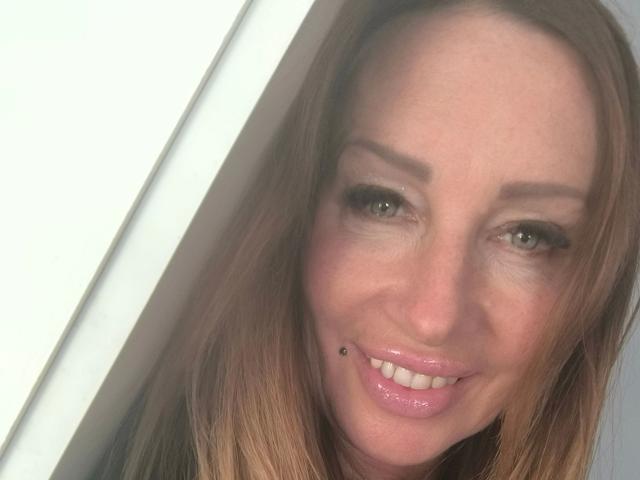 MissJenn-hot has a cute face with light brown hair, green eyes, and a small black mole on her bottom lip. She's smiling with shiny pink lips.