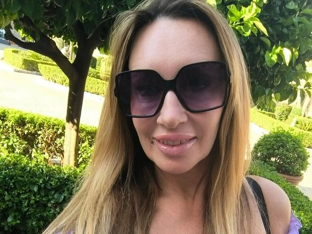MissJenn-hot with blonde hair, wearing black sunglasses, and a purple top, showing a slight pout with glossy pink lips, standing outdoors under a tree.